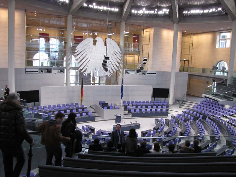 zittingzaal bundestag.jpg - Onze rondleiding stopt in de zittingszaal. De architect heeft getracht om de Duitse adelaar in deze zaal een slanker uiterlijk te geven. Daarvoor diende hij meer als tachtig ontwerpen in, die allemaal door het parlement afgewezen werden. Men besloot om een vergrote versie van de adelaar die al in Bonn hing te gebruiken. Aangezien de adelaar voor een glazen wand hangt, is op de achterzijde een slankere en kleinere versie te zien, zoals de architect graag wilde.
